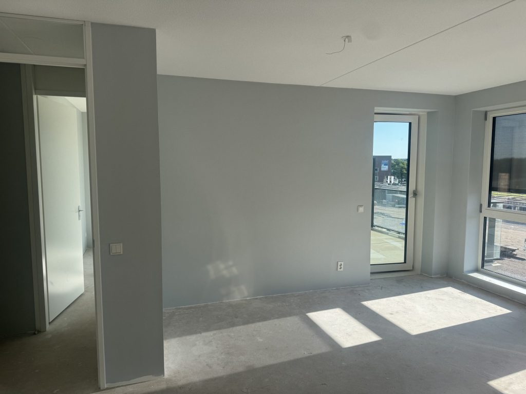 Nieuw appartement met lichtgrijze wanden, een balkondeur en een betonvloer, net na het afwerken